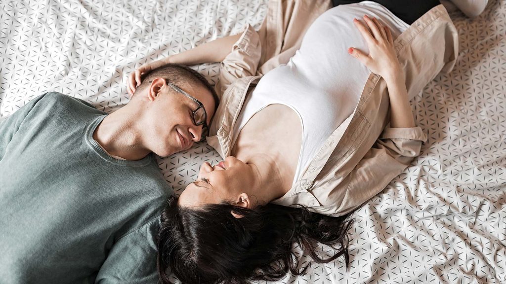 Schwangeres Paar liegt auf dem Bett und tauscht Zärtlichkeiten aus.