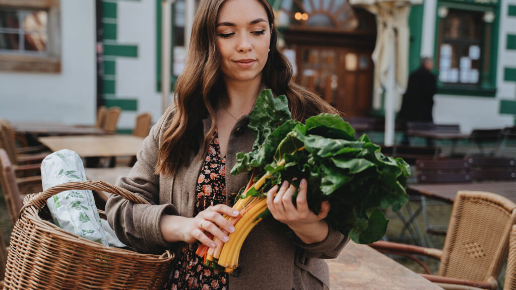 Schwangere Frau mit Mangold in der Hand und Einkaufskorb unter Arm mit veganen Produkten